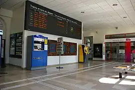 Hall d'accueil vide avec le panneau d'indication des trains sur le mur et les automates en-dessous. L'accès aux guichets est au fond.