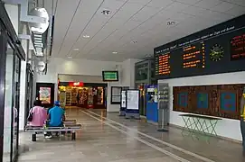 Hall d'accueil avec le panneau d'indication des trains sur le mur et les automates en-dessous. Un commerce vendant des journaux est situé au fond.
