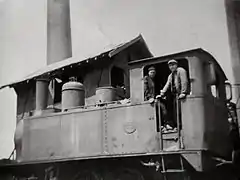 Photo noir et blanc d'une petite locomotive à vapeur vue de trois-quarts arrière avec son chauffeur et son mécanicien.