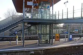 Cliché couleur. Au-dessus de voies de chemin de fer, une passerelle est desservie par deux escaliers et deux ascenseurs.