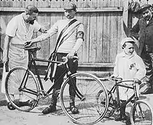 Photographie en noir et blanc d'un homme portant son vélo et un petit garçon avec une petite bicyclette, entourés de deux autres hommes.