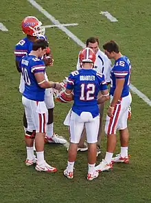 Quatre joueurs de football américain en maillot bleu discutant avec un entraîneur sur un terrain de football américain.