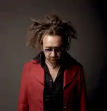 Portrait d'un homme d'une soixantaine d'années portant les cheveux longs en dreadlocks et un bouc, avec une veste rouge, une chemise noire et des lunettes de soleil.