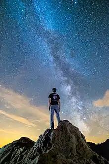 De nuit un homme se tient debout, de dos, regardant vers le ciel ; la Voie Lactée s'éparpille devant lui en une longue traînée brillante par des étoiles bleues et blanches.