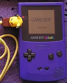Photo d'une Game Boy mauve avec un câble link jaune en forme de Pikachu.