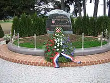 Un momument commémoratif, avec des inscriptions en serbo-croate et en allemand à la mémoire de la "tragédie de Bleiburg" ; devant le momument, une gerbe de fleurs aux couleurs du drapeau croate.