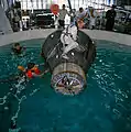 Photographie en couleur de Stafford et Shirra dans une piscine avec une reproduction du vaisseau Gemini.