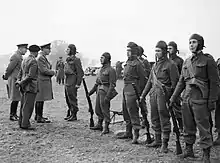 Photographie en noir et blanc de sept hommes vêtus de combinaisons sombres, se tenant devant trois officiers