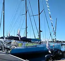 Vu de trois quarts arrière, voilier bleu amarré au ponton. Rouf et casquette jaune et blanc.