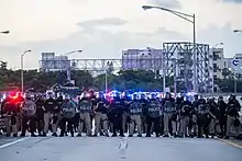 Plusieurs policiers armés en uniforme avec casques et boucliers sur une autoroute barrée, plusieurs édifices et des espaces verts en arrière-plan.