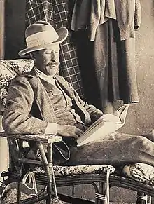Photographie sépia d'un homme lisant assis dans un fauteuil et portant un costume complet ainsi qu'un chapeau.