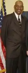 George Gervin en 2008, debout en costume, d'un drapeau américain derrière lui.