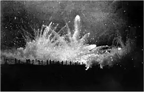 Photo noir et blanc d'un tir de nuit, gerbe de feu éclairant des barbelés.