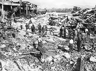 Hommes travaillant parmi des débris de bombardement, avec des restes de bâtiments sur la gauche.