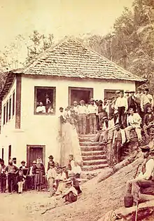 Photo montrant un groupe de personnes rassemblées autour et sur les marches d’une maison en crépi blanc avec un toit couvert de bardeaux de bois située sur le versant abrupt d’une colline boisée