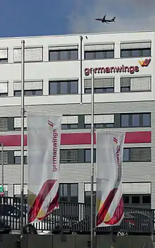 Photo de deux drapeaux abaissés aux couleurs blanches, oranges et rouges, en face d'un bâtiment blanc.