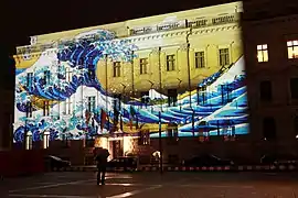 Photo couleur (prise de vue nocturne) de la façade d'un bâtiment en pierre sur laquelle est projetée une vidéo représentant une estampe figurant une très haute vague déferlant sur de longues barques conduites par des hommes.