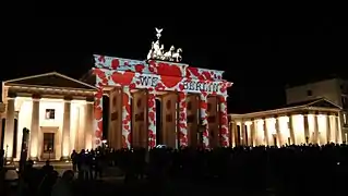 Photo couleur (prise de vue nocturne) du fronton d'une porte en pierre soutenu par six colonnes. L'ensemble est illuminé (des cœurs rouges sur fond blanc, avec le message en noir : « We ♥ Berlin »). Au premier plan, une foule de badauds.