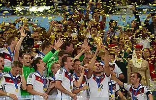 Joueurs de football en tenue claire, sous des confettis, fêtant dans la tribune avec le trophée.