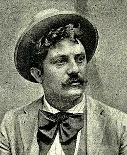 Image en noir et blanc d'un buste d'homme position trois quart, tourné vers la droite, cheveux frisés dépassant sous un chapeau, avec moustache, flocon noir au col.