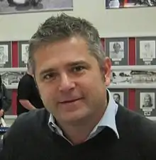 Photographie d'un homme blanc, vu de face, inclinant sa tête, cheveux gris, souriant.