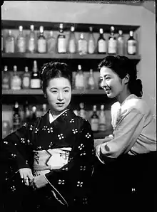 Une femme en kimono sombre (Kinuyo Tanaka) se tient de face au comptoir d'un bar. À droite, une jeune femme en chemisier blanc (Kyōko Kagawa) la regarde en souriant. Dans le fond, plusieurs étagères supportent des rangées de bouteilles (image en noir et blanc).