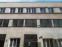 Photographie d'un bâtiment à la façade beige, aux fenêtres rectangulaires ; en bas à droite de l'image, une plaque avec un texte et une représentation artistique.