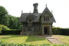 Photo de la façade d'une maison à l'architecture fantasque, avec un toit ouvragé, entourée d'une pelouse tondue à ras et d'une haie