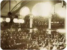 Une photo montrant un grand nombre d'hommes assis sur des gradins en demi-cercle dans une salle voûtée alors qu'une grande foule regarde d'un balcon à arcades