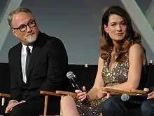 Photographie d'un homme en costume et d'une femme en robe assis devant un écran de projection. Celle-ci tient un micro dans sa main et regarde avec malice l'objectif de l'appareil photo.