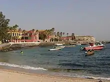 Photographie d'une petite plage bordée de palmiers et de maisons, avec un quai de pierre et quelques embarcations légères au mouillage.