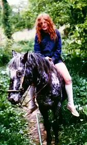 Dans un chemin arboré, une jeune fille rousse, sans casque, monte un poney gris à cru.