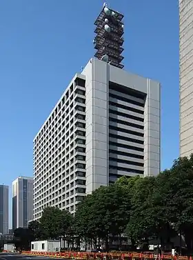 Photo d'un immeuble de bureaux d'une vingtaine d'étages, vu de 3/4 droit. La rue au pied de l'immeuble est bordée par une rangée d'arbres. L'mmeuble est surmonté d'une tour d'antennes de sept étages