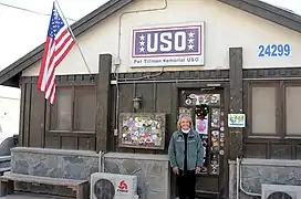 Une femme se tient devant l'entrée d'un bâtiment sur lequel il y a une pancarte « USO - Pat Tillman Memorial USO » et un drapeau américain.