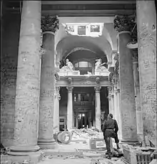 Photo noir et blanc, prise le 3 juillet 1945 à Berlin, montrant l’intérieur d’un hall du Reichstag en ruines. Au premier plan, un soldat, debout et de dos, regarde les innombrables graffitis écrits sur les colonnes de couleur claire du lieu.