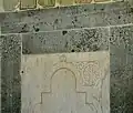 Vue d'un panneau en marbre blanc, bordé de bandeaux verts, qui se trouve à gauche de la niche. Il est sculpté d'un arc recti-curviligne, dont l'un des écoinçons est orné d'une inscription rayonnante.