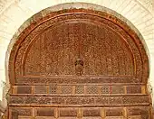 Photographie de la partie supérieure de la grande porte centrale de la salle de prière. Le décor sculpté, notamment celui du tympan, comprend des éléments végétaux, géométriques et épigraphiques.