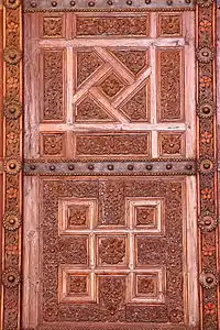 Photographie d'un vantail en bois sculpté de la grande porte de la salle de prière. Des bandes de tôle, ornées de clous à tête ronde, séparent les panneaux ouvragés de chaque vantail.