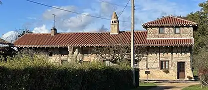 Un long bâtiment à colombages rempli de briques surmonté d'une cheminée sarrasine elle-même surmontée d'une croix.