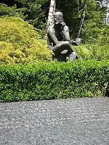 Horizontal gravestone saying "JAMES JOYCE", "NORA BARNACLE JOYCE", GEORGE JOYCE", and "…ASTA OSTERWALDER JO…", all with dates. Behind the stone is a green hedge and a seated statue of Joyce holding a book and pondering.