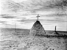 Photographie noir et blanc d'un monticule de neige, surmonté d'une croix.