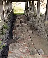 Photo d'une allée couverte encadrée par deux murs de pierre bas. Le sol est partiellement dallé, avec trois petites stèles rectangulaires alignées sur la droite