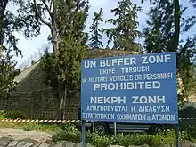Photo d'un panneau sur lequel il est écrit : zone tampon des Nations unies, en langue anglais et grecque. En arrière-plan, un mur fortifié et surmonté d'un grillage.