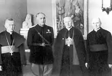 Un homme en uniforme noir, en compagnie de trois ecclésiastiques en habit.