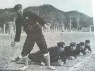 Photo noir et blanc d'un jeune homme, au premier plan à gauche, en tenue militaire sombre, sur le point de lancer une grenade. Quatre autres jeunes hommes sont allongés sur le sol derrière lui.