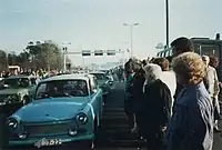 Voitures Trabants est-allemandes roulant au milieu d'une foule dense. Des portiques métalliques et une tour d'observation sont visibles à l'arrière-plan.