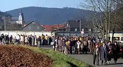 Un grand nombre de personnes de tous ages se trouvent le long d'une route en face d'un mur de béton derrière lequel se trouvent des maisons et une église.