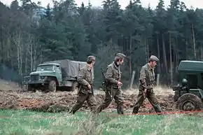 Trois hommes en uniforme et armés de fusils d'assauts marchent en colonne devant une bande de terre labourée. Deux camions se trouvent à l'arrière-plan devant une foret dense.