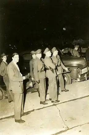 Photographie noir et blanc de six policiers, plusieurs d'entre eux matraque à la main, et d'un homme en habit cravate.