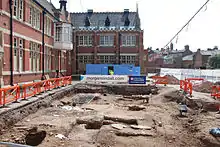 Photo panoramique d'une section du sol déblayée, entourée de barrières de sécurité orange. Des bâtiments se dressent sur le côté gauche.